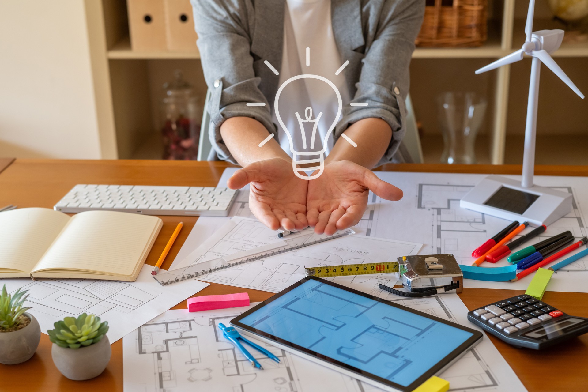 Architect presenting innovative project for sustainable building with renewable energy. Showing light bulb icon above open hands. Surrounded by blueprints. Tablet. Wind turbine model and solar panel